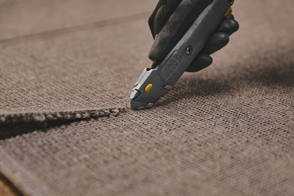 Photo of STANLEY 10-499 utility knife cutting brown carpet, close-up view of blade in use.