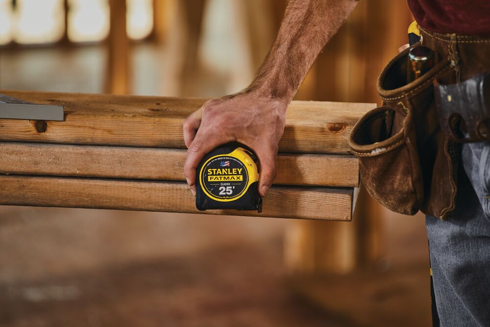 Photo of STANLEY 33-725 tape measure held by a person measuring wooden beams in a construction setting.