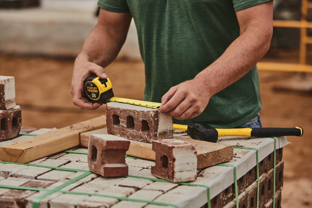 STANLEY 33-725 tape measure in use measuring a brick at a construction site, bricks and hammer visible on table.