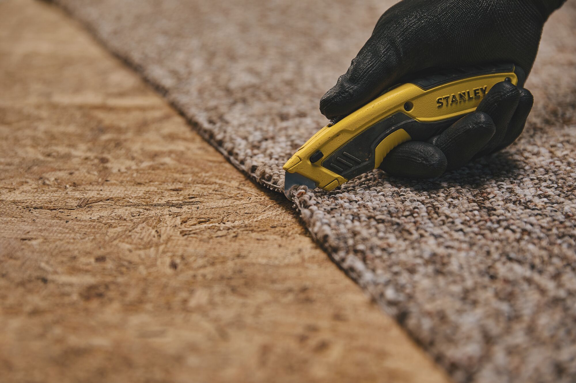 Stanley STHT10479 utility knife cutting carpet, close-up, user wearing black gloves.