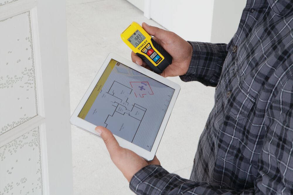 STANLEY STHT77343 laser distance measurer held with tablet displaying floor plan, indoor setting.